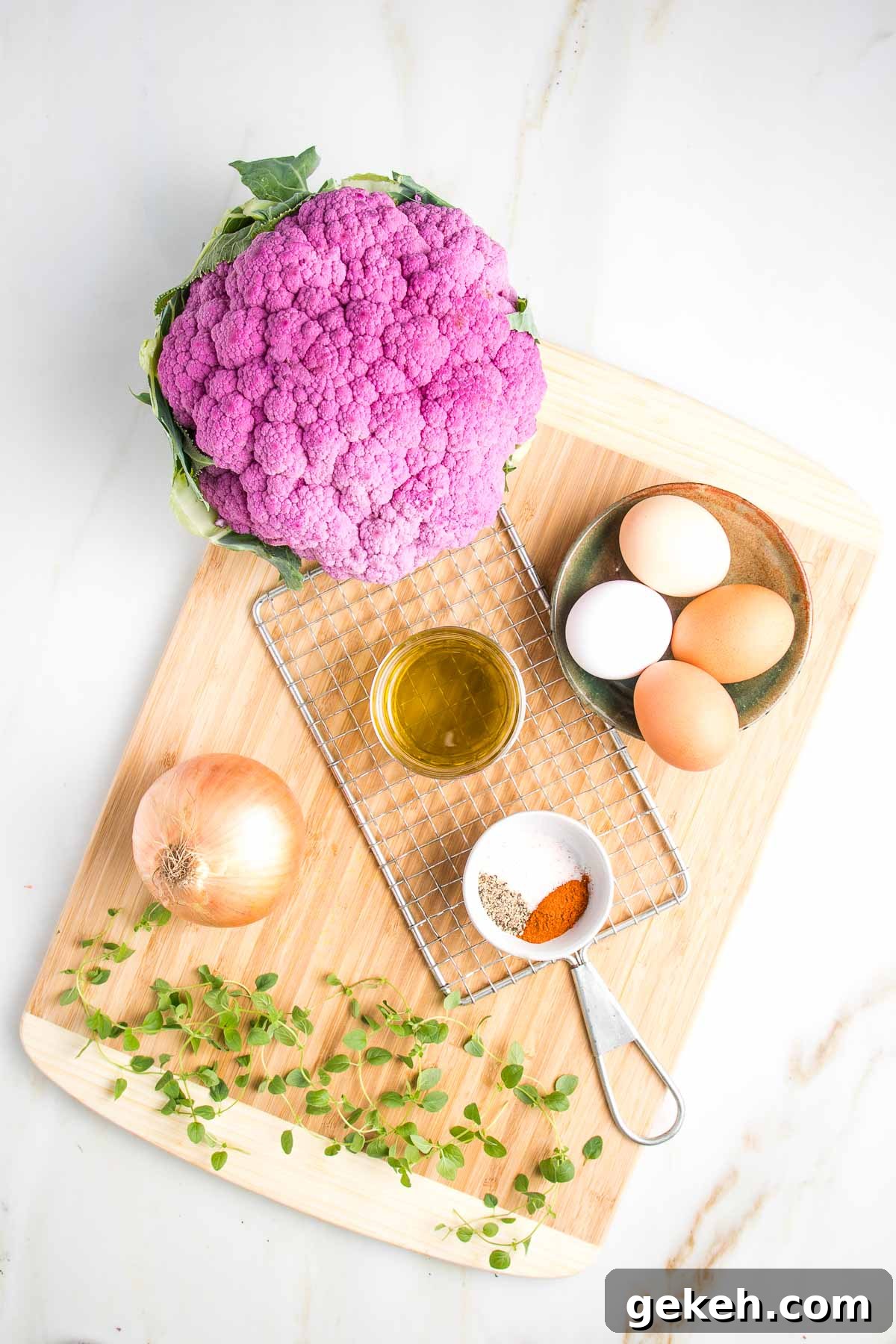 Cauliflower kugel recipe ingredients on a cutting board:  large head purple cauliflower, eggs, fresh oregano, spices, olive oil, onion and shredder.