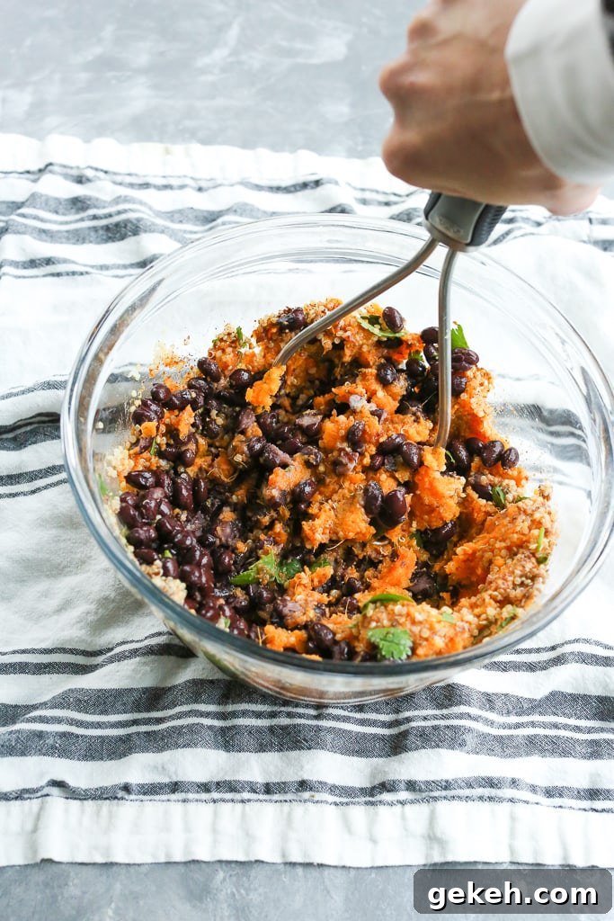 Sweet Potato and Black Bean Burger Recipe for Flavorful Veggie Patties 5 cooked sweet potato, black beans, quinoa, cilantro and spices in a glass bowl with a hand holding a potato masher