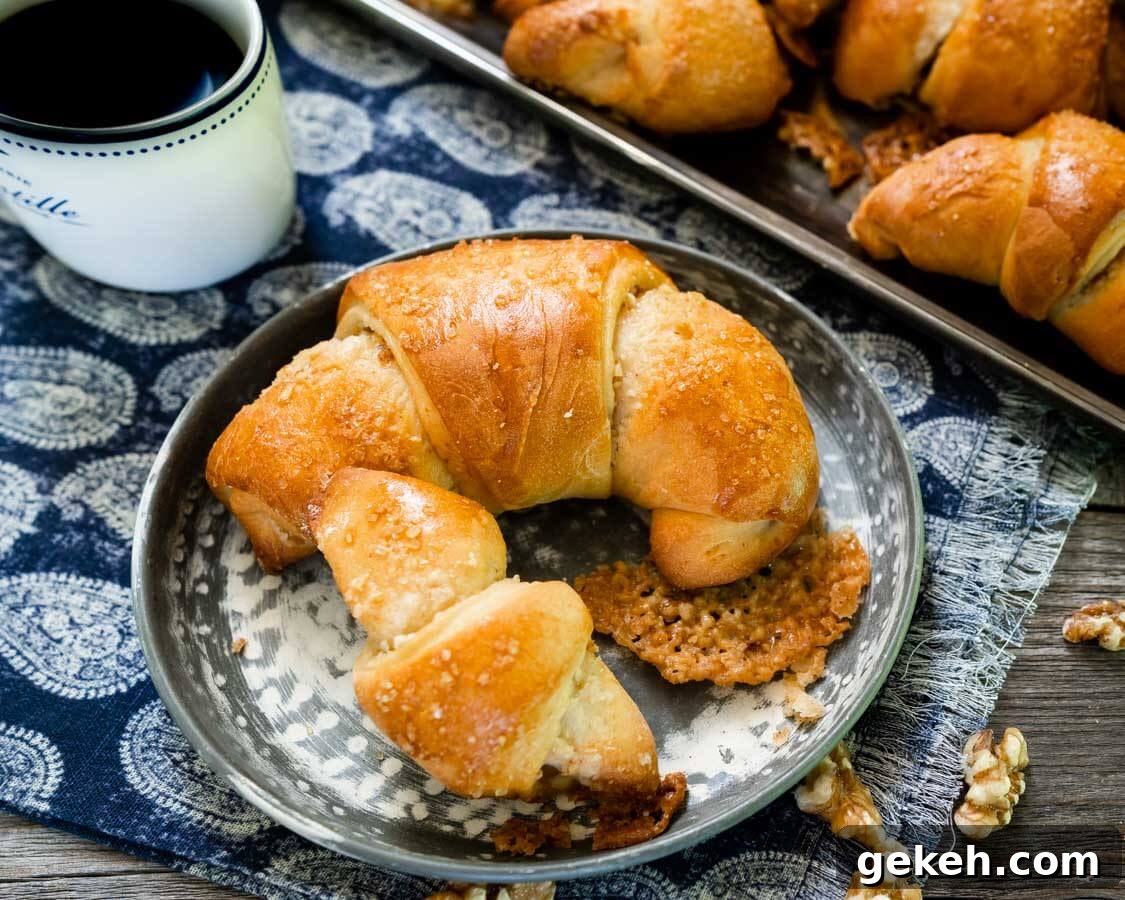 Authentic Kifli (Hungarian Walnut Roll) Recipe for Homemade Pastry 2 A metal plate holding a large and small Kifli Walnut roll sits on a blue paisley napkin. A tray of Kifli rolls and a cup of coffee sit in the back.