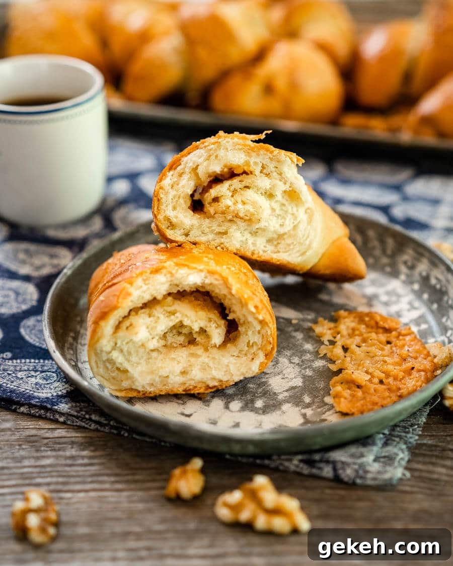 Authentic Kifli (Hungarian Walnut Roll) Recipe for Homemade Pastry 4 A Kifli Walnut Roll cut in half showing the cinnamon swirl inside. A cup of coffee and a tray of whole Kifli rolls sit in the background.