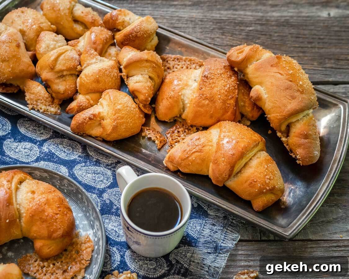 Authentic Kifli (Hungarian Walnut Roll) Recipe for Homemade Pastry 5 A silver tray of Kifli Walnut Rolls sitting on a blue napkin over a wooden board. A cup of coffee and a partial plate with a Kifli roll sit in the foreground.