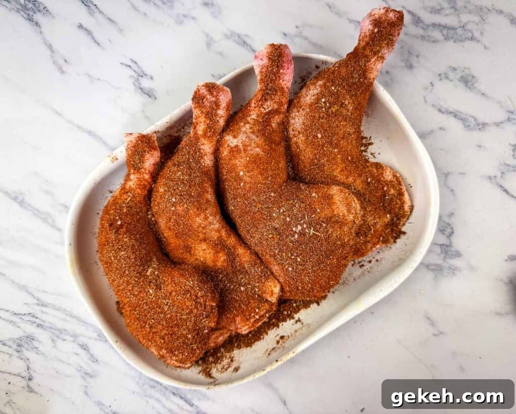 Four raw chicken leg quarters coated with a reddish spice rub are arranged on a white oval dish on a marble surface.