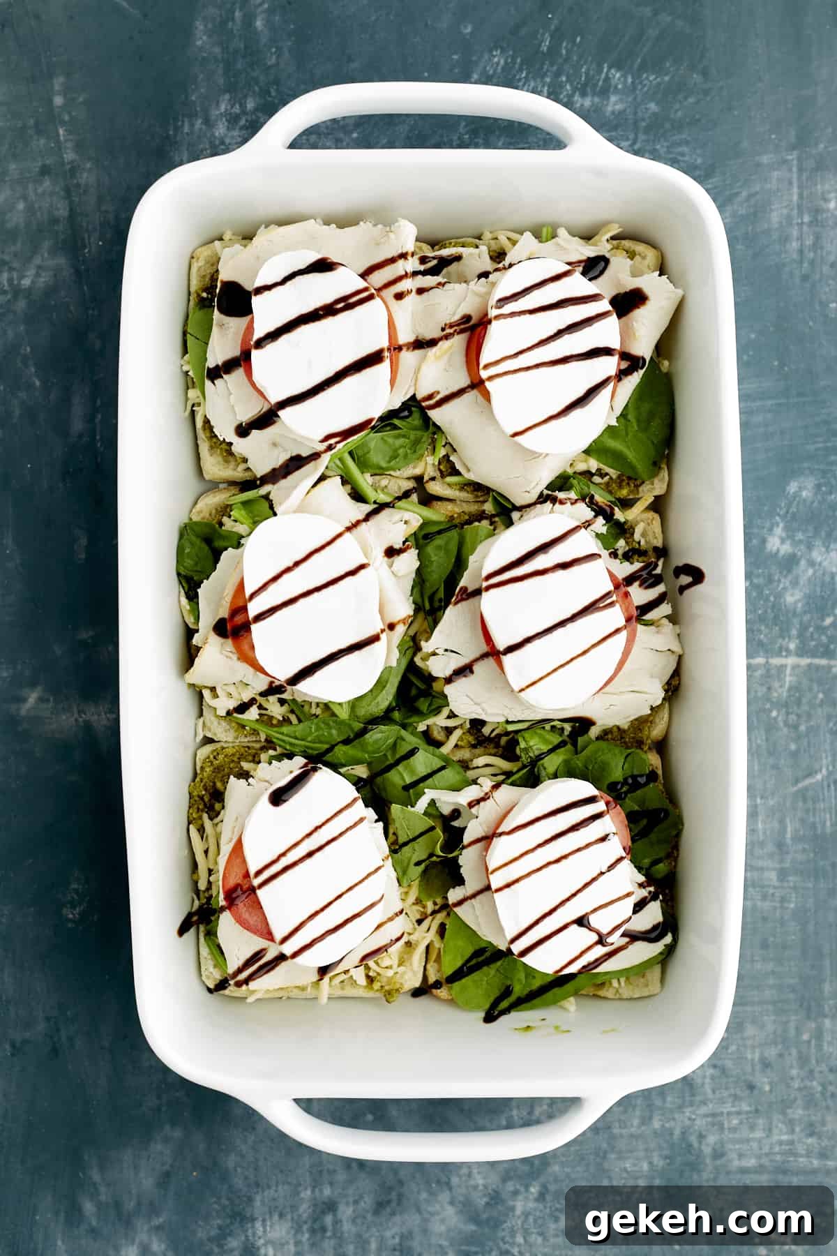 Turkey Caprese Sandwich with Fresh Mozzarella & Basil 9 sliced ciabatta buns in a white baking dish topped with pesto, spinach, turkey, tomatoes, mozzarella, and balsamic glaze.