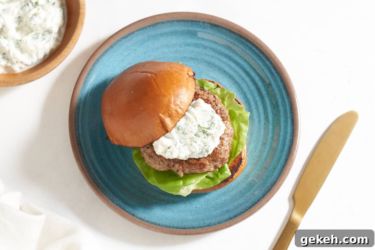 Juicy Ground Lamb Burgers with Creamy Feta Sauce 7 A lamb burger topped with feta yogurt sauce with a bun on a blue plate, a bowl of sauce is in the upper left corner.