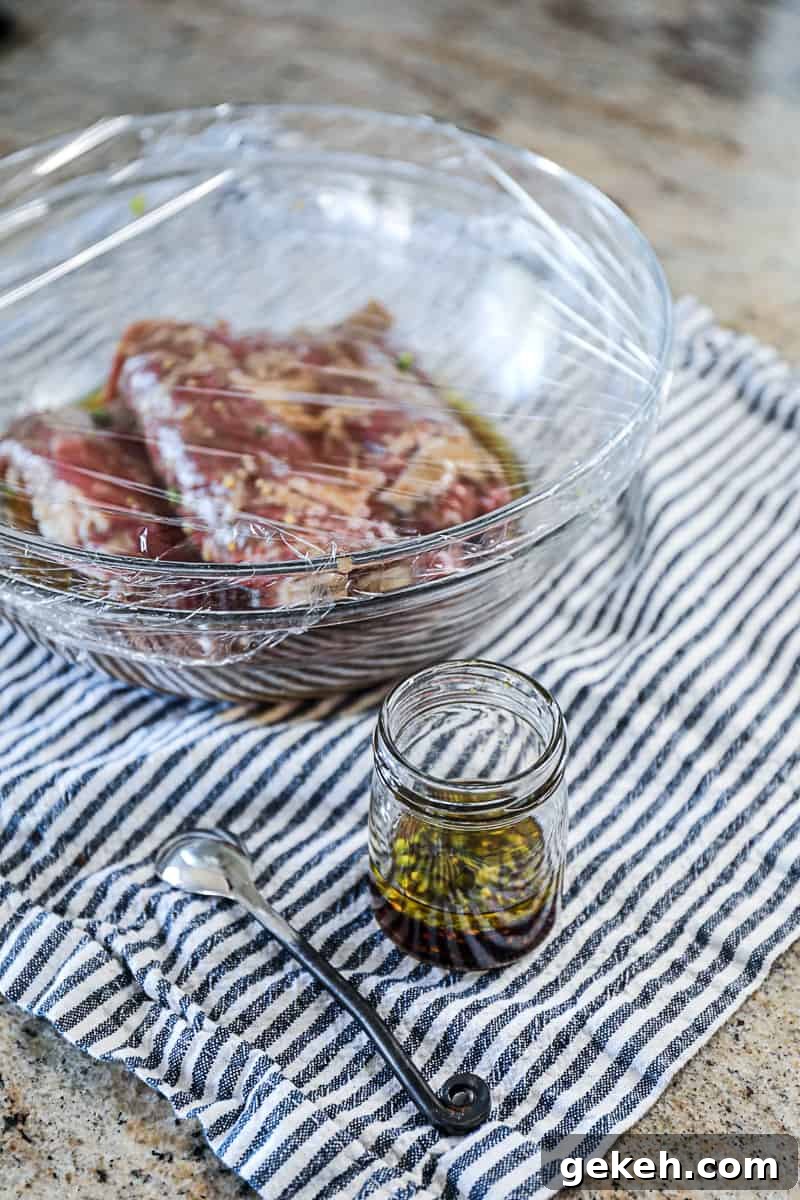 Side shot of skirt steak marinading in a large bowl wrapped in plastic wrap with a side marinade in a jar and a spoon on a counter.