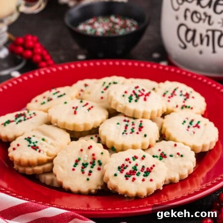 Holiday cookies with scalloped edges topped with red, green, and white sprinkles, arranged on a red plate surrounded by festive decor, including a striped napkin, red berries, and a "Cookies for Santa" jar.