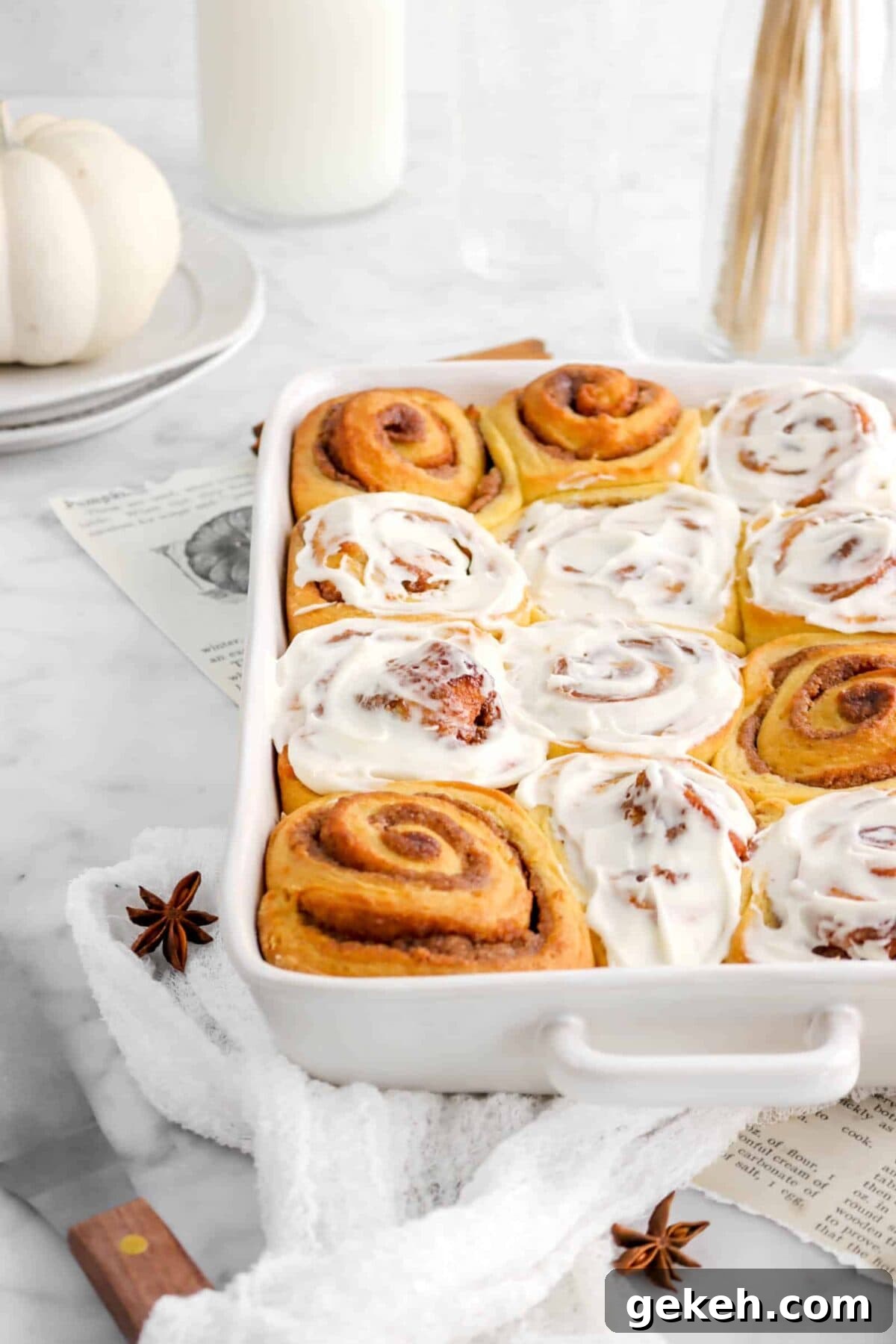 pumpkin cinnamon rolls in white casserole with white cheese cloth, plates, and star anise pods