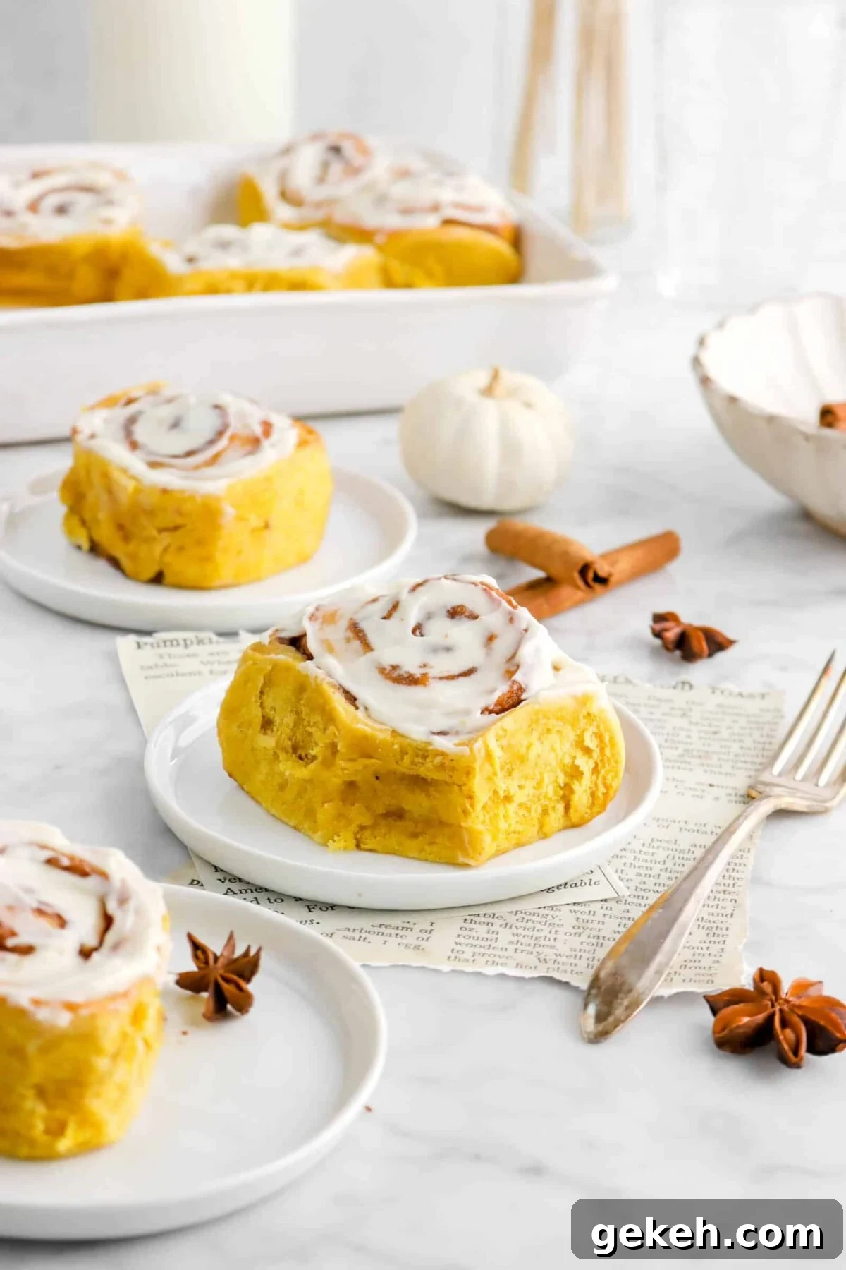 pumpkin cinnamon rolls on marble counter with three on three white plates, with tray of pumpkin rolls behind, stat anise pods, cinnamon sticks, and a fork