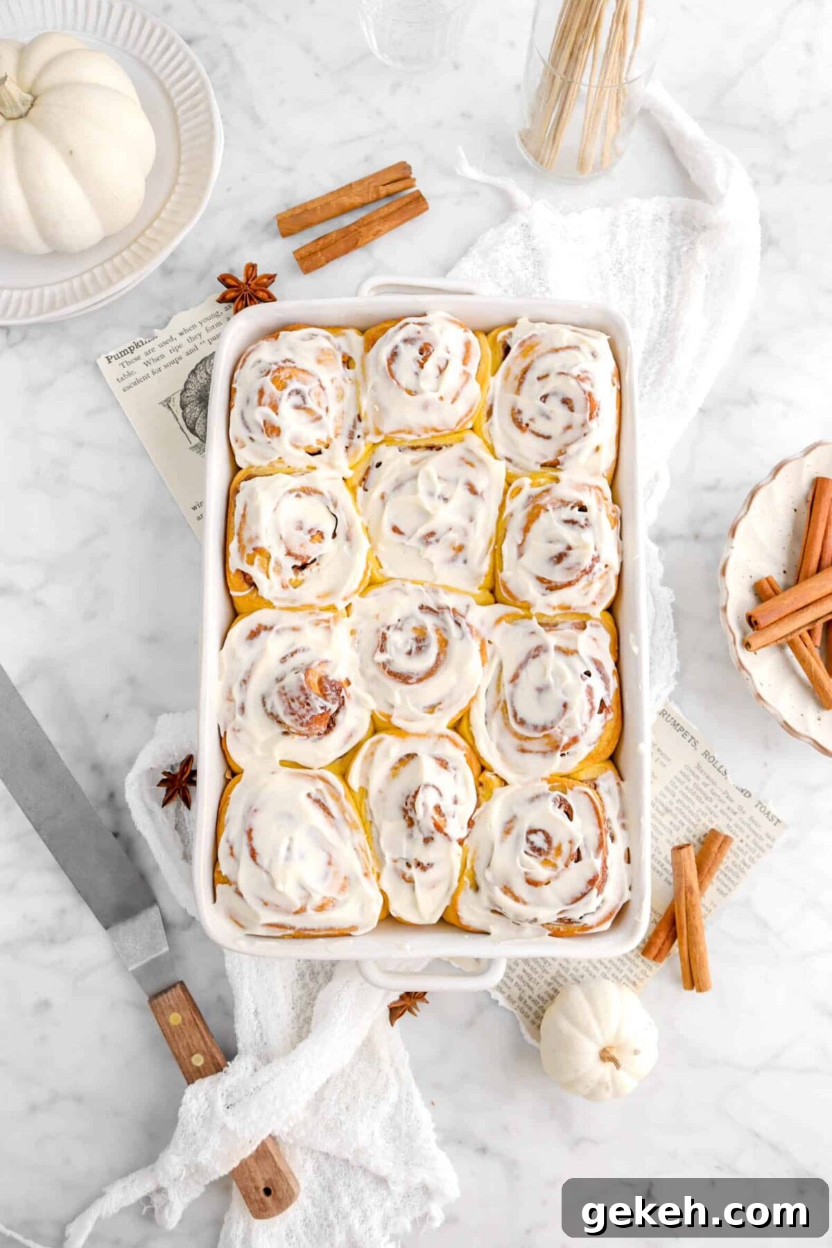 frosted pumpkin cinnamon rolls on marble counter with cinnamon sticks, white cheese cloth, and book pages