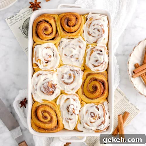 overhead shot of pumpkin cinnamon rolls on white cheese cloth, cinnamon sticks, white pumpkins, and icing spatula