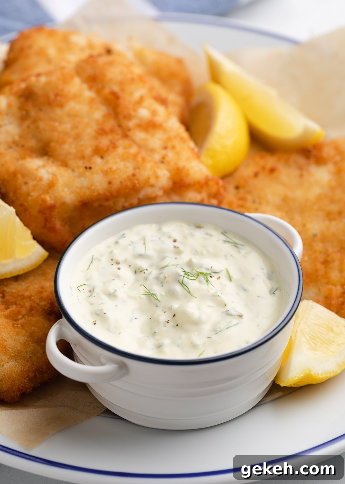homemade tartar sauce in a white handled bowl with blue rim on a platter of fried fish with lemon wedges