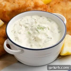 homemade tartar sauce in a white handled bowl with blue rim on a platter of fried fish with lemon wedges
