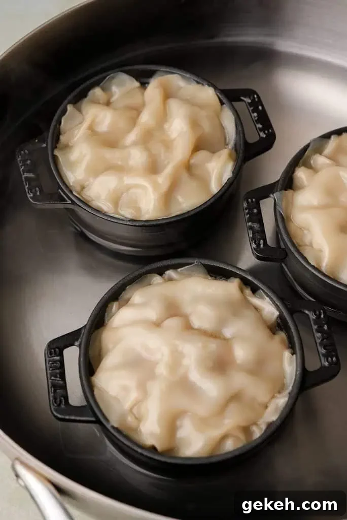 Three mini black cocottes filled with layered wonton wrappers sit inside a large pot with water, showing the dumplings mid-steam before cooking is finished.