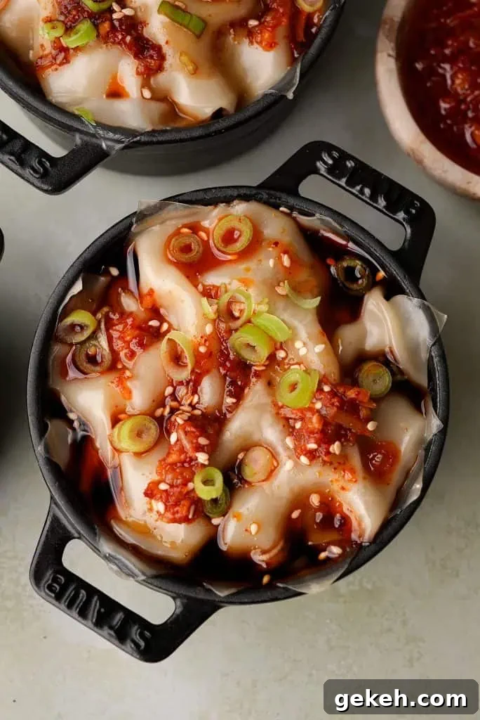 A top-down view of a mini cocotte shows steamed dumpling lasagna topped with chili oil, sliced scallions, and sesame seeds, with a bowl of chili oil partially visible nearby.