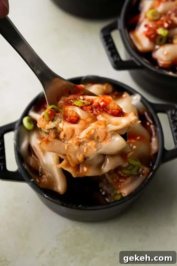 A spoon lifts a layered portion of dumpling lasagna from a mini cocotte, revealing the tender pork filling and softened wonton wrappers coated in chili oil.