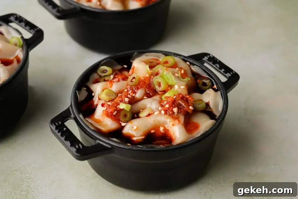 A small black cocotte filled with layered wonton wrappers is topped with chili oil, sliced scallions, and sesame seeds on a light stone surface.