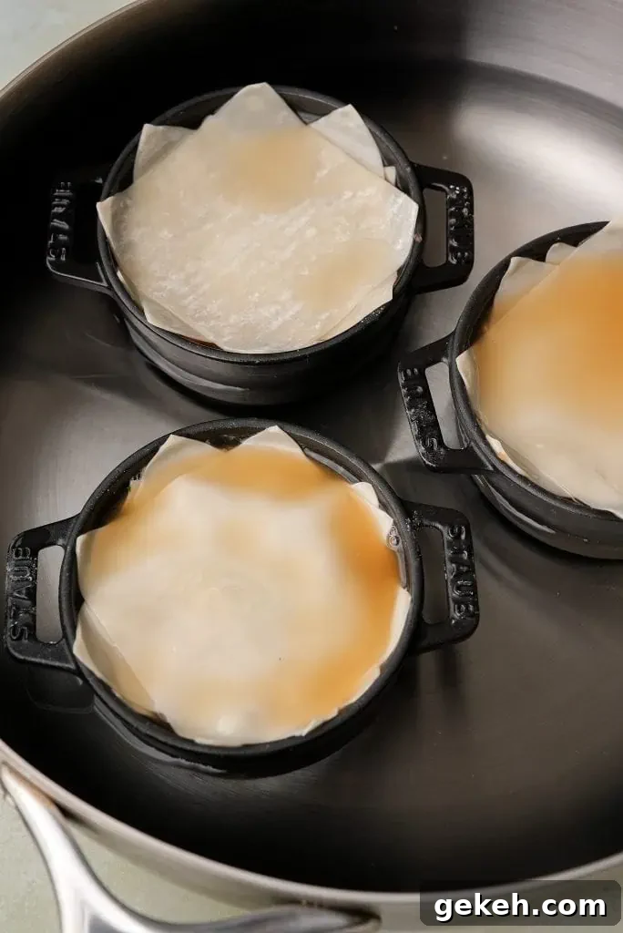 Mini cocottes layered with wonton wrappers and chicken stock sit inside a large pot filled with water, set up for steaming on the stovetop.