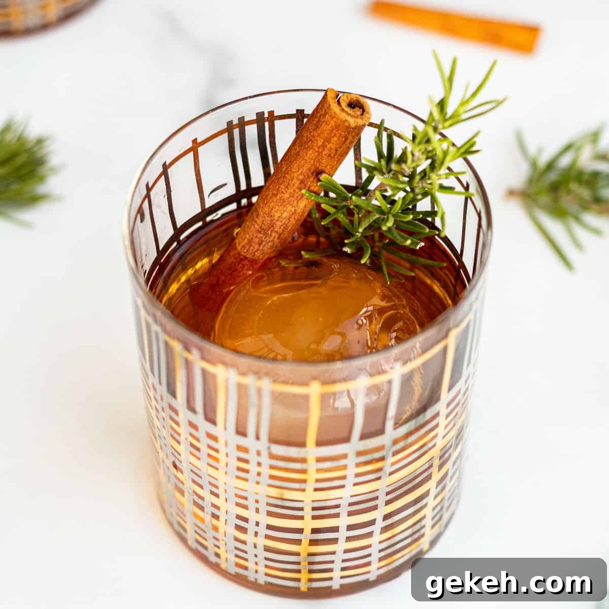 cocktail glass with a clear ice ball and winter old fashioned garnished with rosemary and cinnamon stick sitting on marble counter
