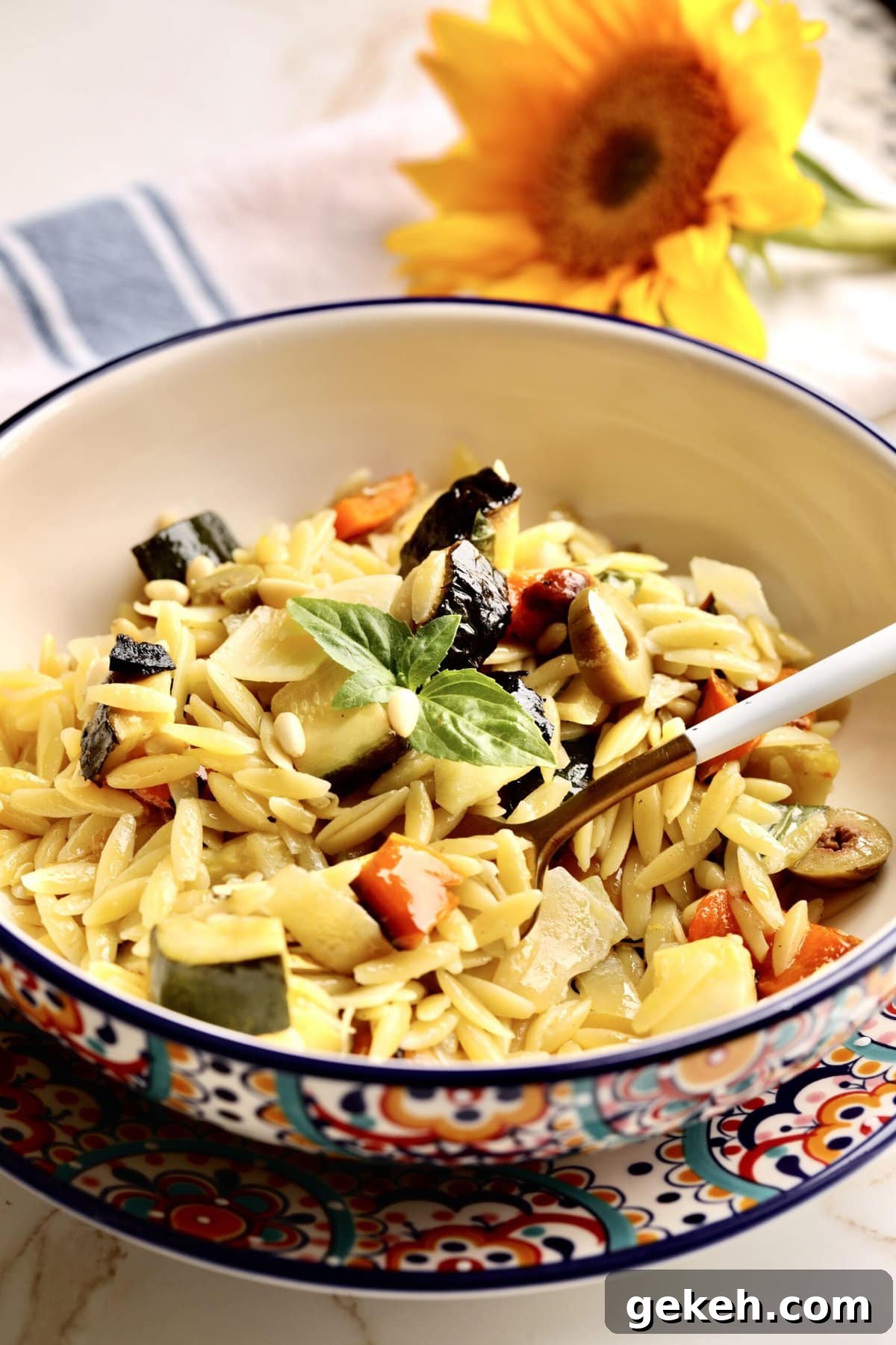 Roasted Vegetable and Parmesan Orzo Recipe 11 big bowl of orzo pasta with roasted vegetables with a fork ready to eat. Flowers in the background.
