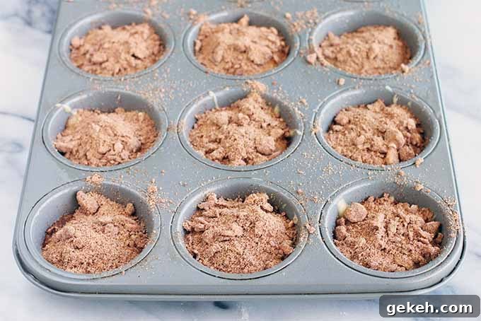 Classic Old-Fashioned Crumb Cake Recipe: Timeless Coffee Cake Delight 13 Nine wells of a metal baking pan are filled with batter topped with a brown cinnamon crumble topping, on a white and gray marble slab.