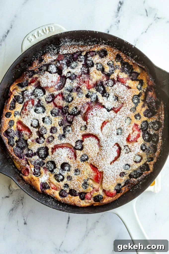 Berry Dutch Baby Pancake: Fluffy Skillet Dessert with Fresh Berries 4 blueberry and strawberry dutch baby in a white skillet and dusted with powdered sugar