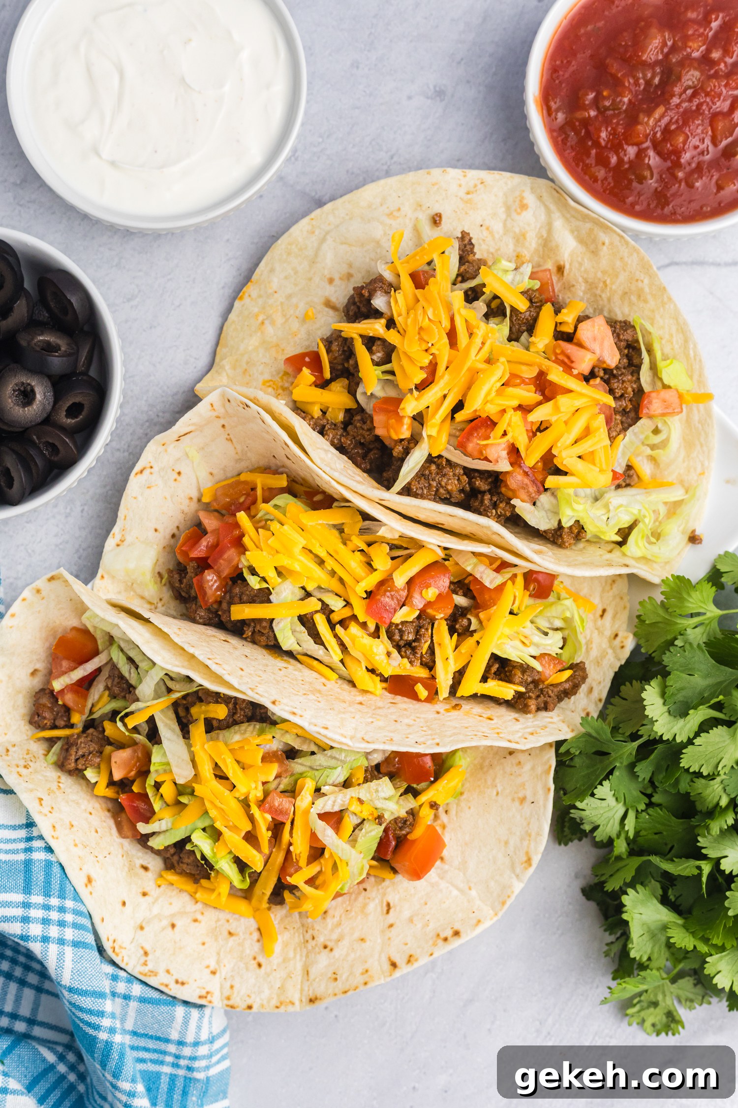 Blackstone Taco Recipe: Crispy Griddle Tacos with Charred Veggies 2 Three soft flour tacos filled with ground beef, lettuce, diced tomatoes, and cheddar cheese, displayed on a white surface with bowls of sour cream, salsa, and black olives nearby.