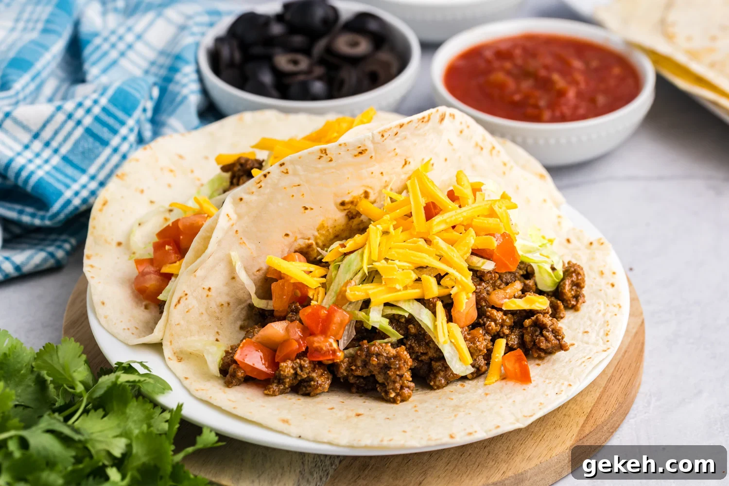 Blackstone Taco Recipe: Crispy Griddle Tacos with Charred Veggies 8 Two soft flour tortillas filled with seasoned ground beef, shredded lettuce, diced tomatoes, and shredded cheddar cheese, served on a white plate with bowls of black olives and salsa in the background.