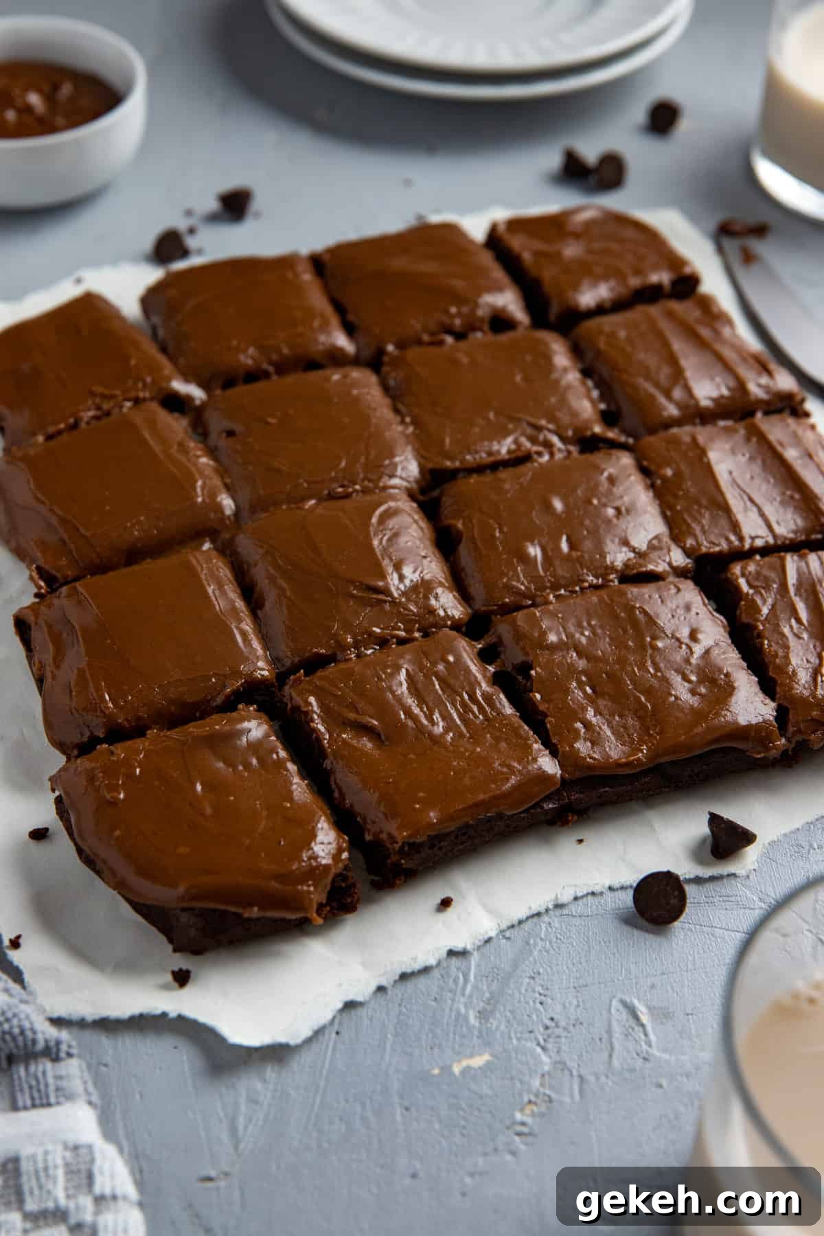 Decadent Dark Chocolate Brownies Recipe for Rich, Fudgy Squares 2 Baked and frosted brownies cut into 16 squares on white parchment paper with brownie crumbs scattered around.