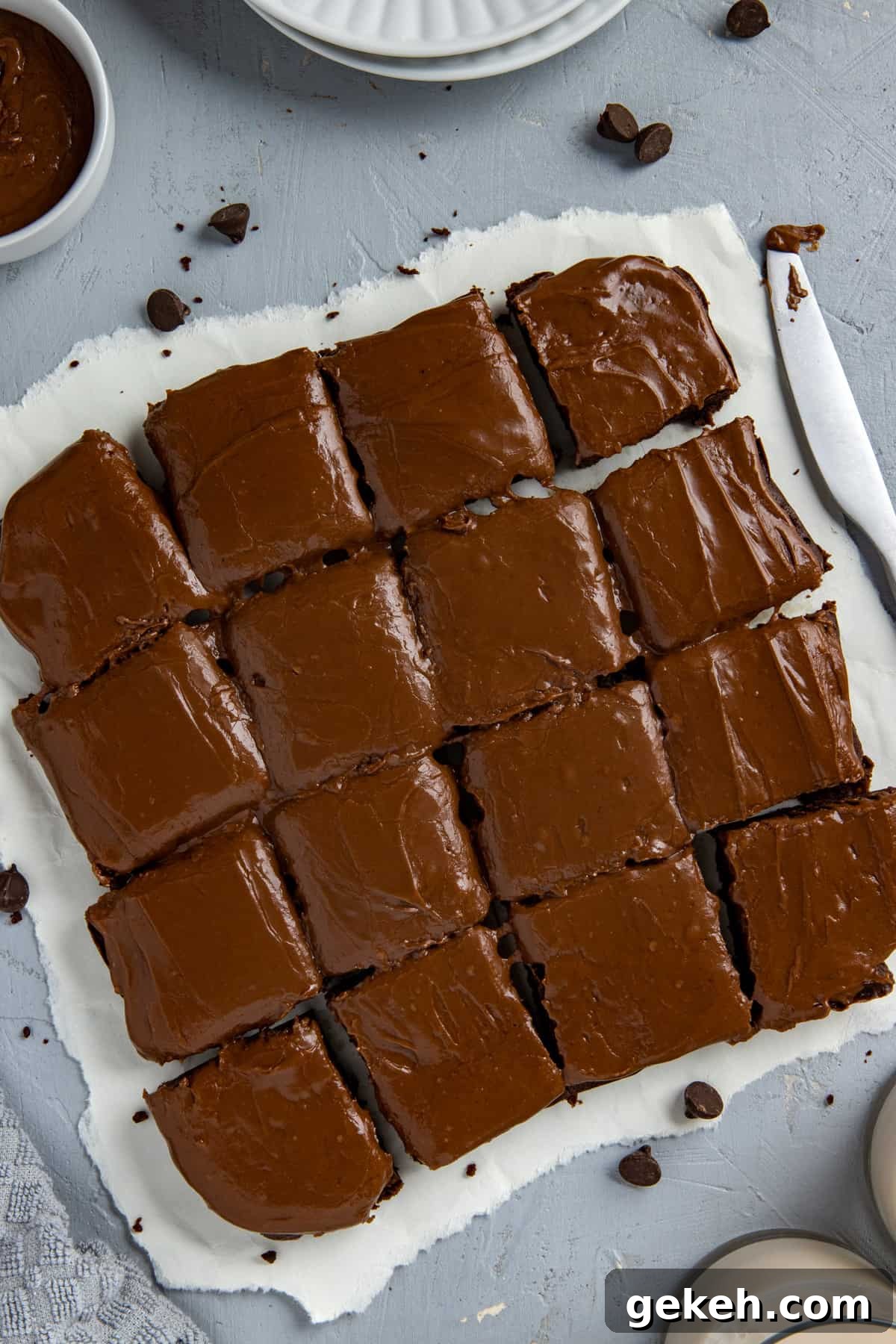 Decadent Dark Chocolate Brownies Recipe for Rich, Fudgy Squares 5 Baked and frosted brownies cut into 16 squares on white parchment paper with brownie crumbs scattered around.