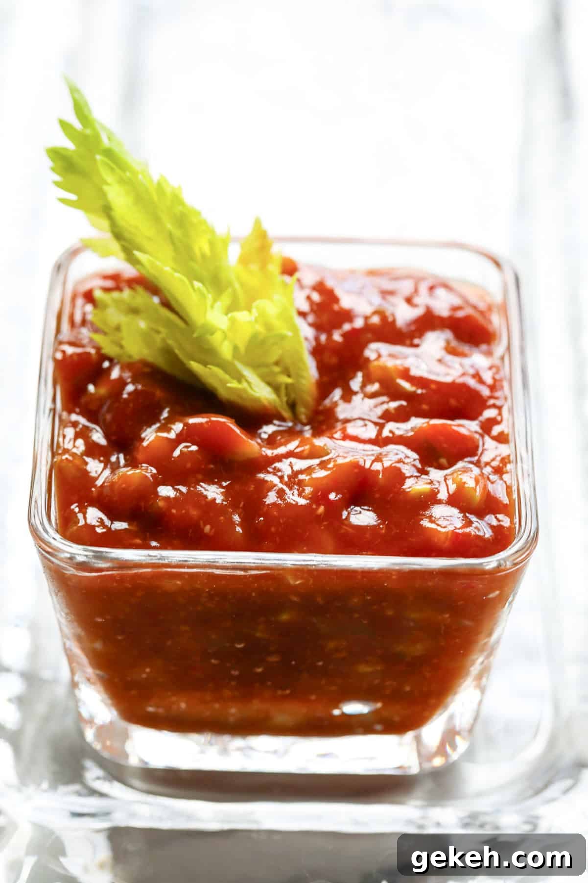 A small square glass bowl filled with homemade cocktail sauce and a celery top garnish to serve with shrimp cocktail.