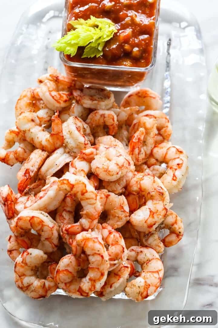 A clear glass platter full of cooked shrimp for shrimp cocktail with cocktail sauce nearby.