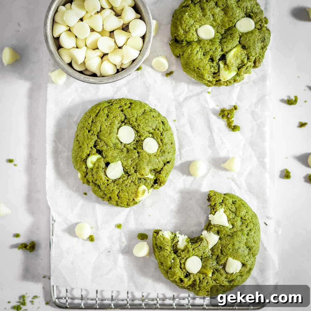 Overhead shot of three matcha cookies, one with a bite taken out on a sheet with parchment paper and a bowl of white chocolate chips on the side.