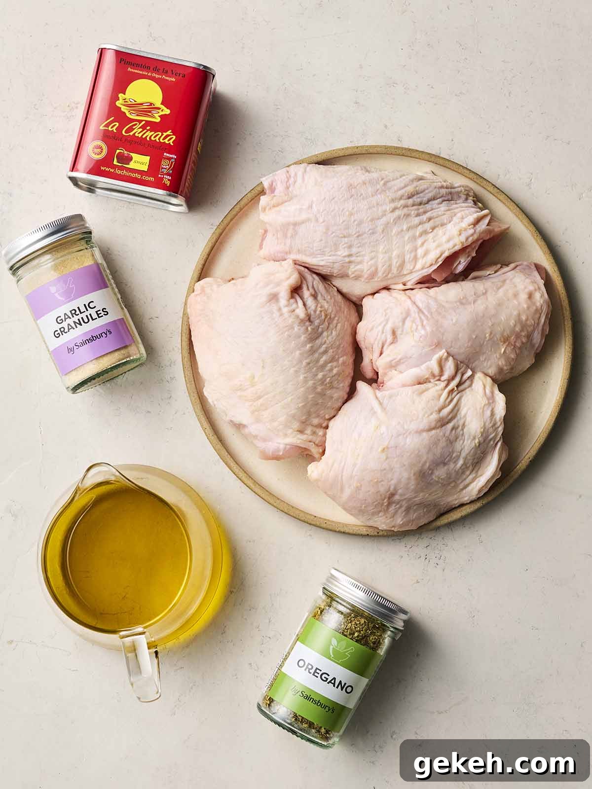 Ingredients laid out on a white counter, including a plate of four chicken thighs with skin on, a jug of oil, and paprika, garlic granules and oregano.