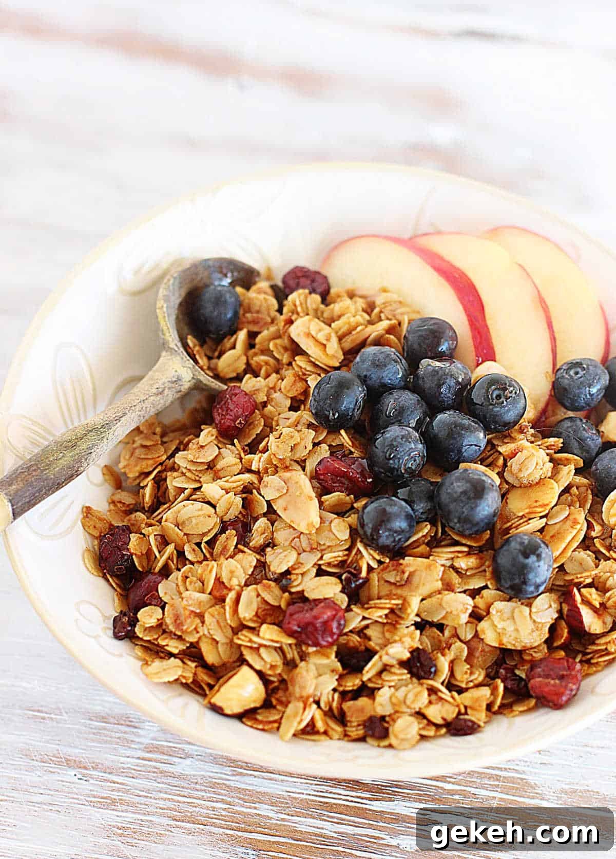 White bowl filled with granola, apples and blueberries, with wooden spoon, on a white table.