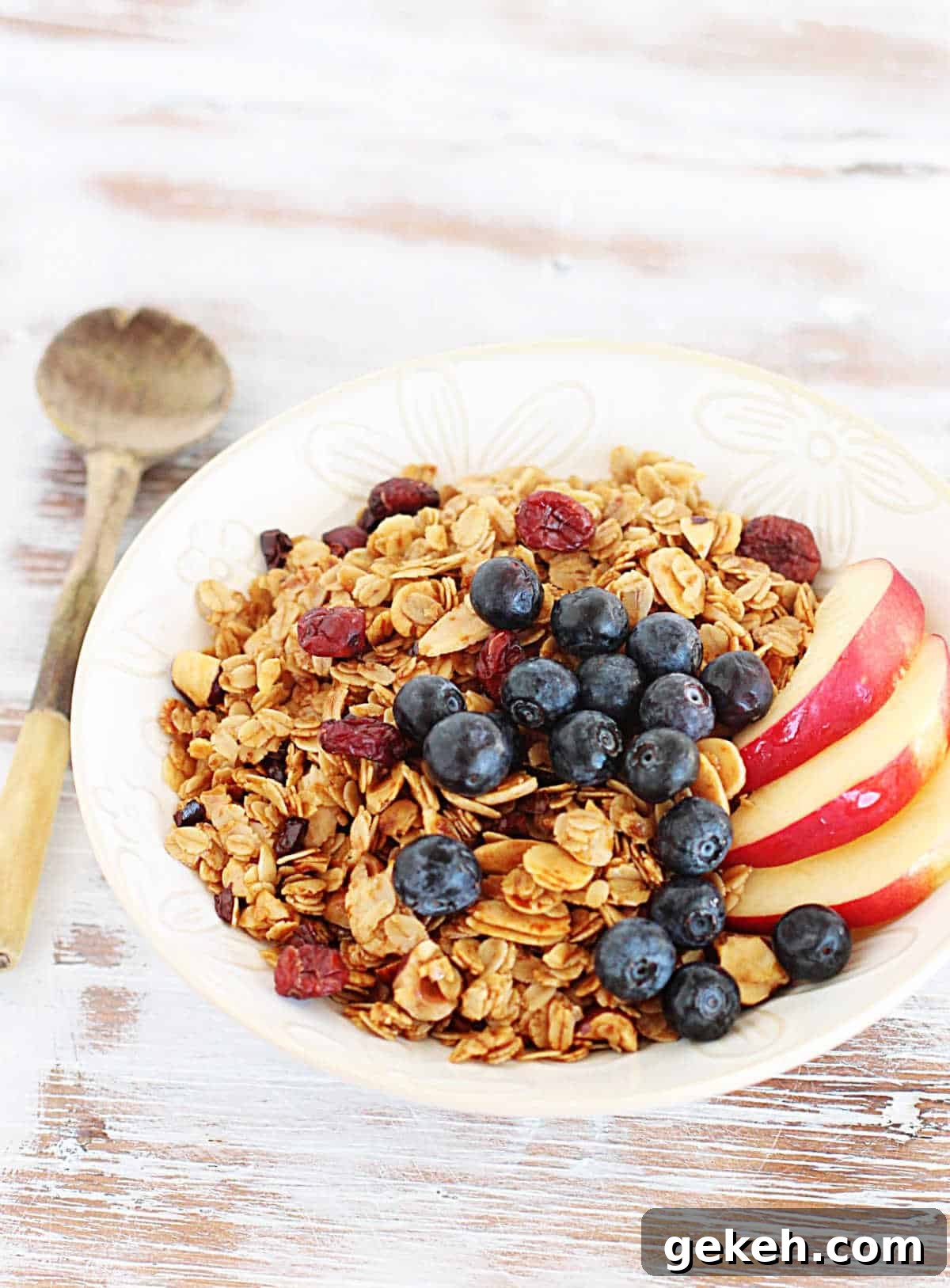 Whitish table with white bowl with granola and fruit, a wooden spoon beside it.