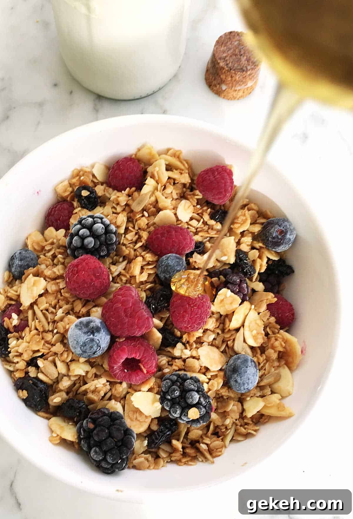 Drizzle of honey pouring onto a white bowl with granola and berries, white surface.