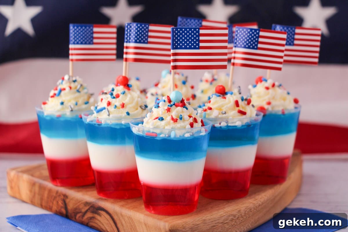 Red white and blue jello shots on a serving board with whipped cream, sprinkles, cocktail stick American flags with American flag in the background.