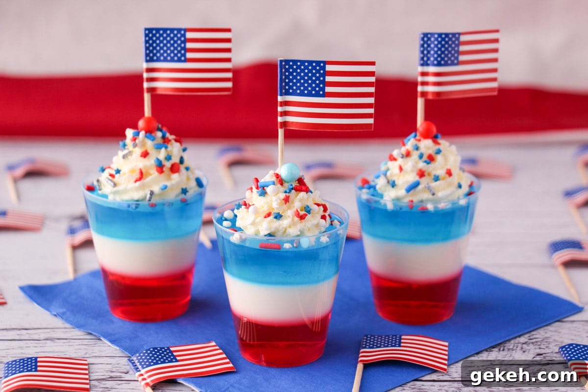 Red white and blue jello shots on blue napkin with whipped cream, sprinkles, cocktail stick American flags, surrounded by cocktail stick American flags.