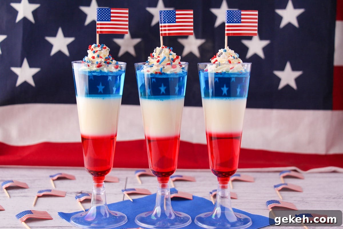 Kid friendly non alcoholic red white and blue jello shots in tall glasses with whipped cream, sprinkles, cocktail stick American flags with American flag in the background.