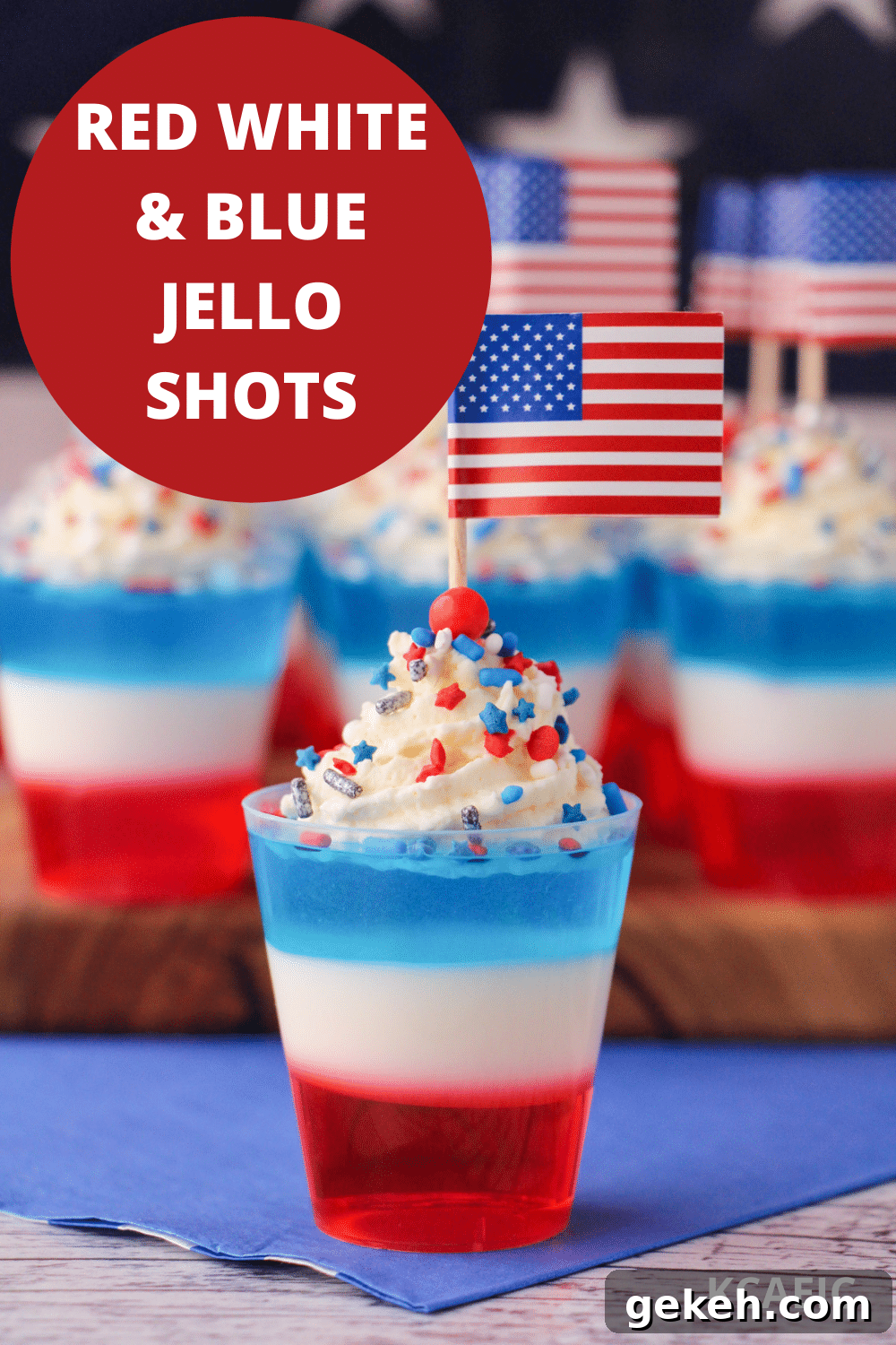 Red white and blue jello shots on a serving board with whipped cream, sprinkles, cocktail stick American flags with American flag in the background, with text overlay, red white and blue jello shots and KCAEIC.