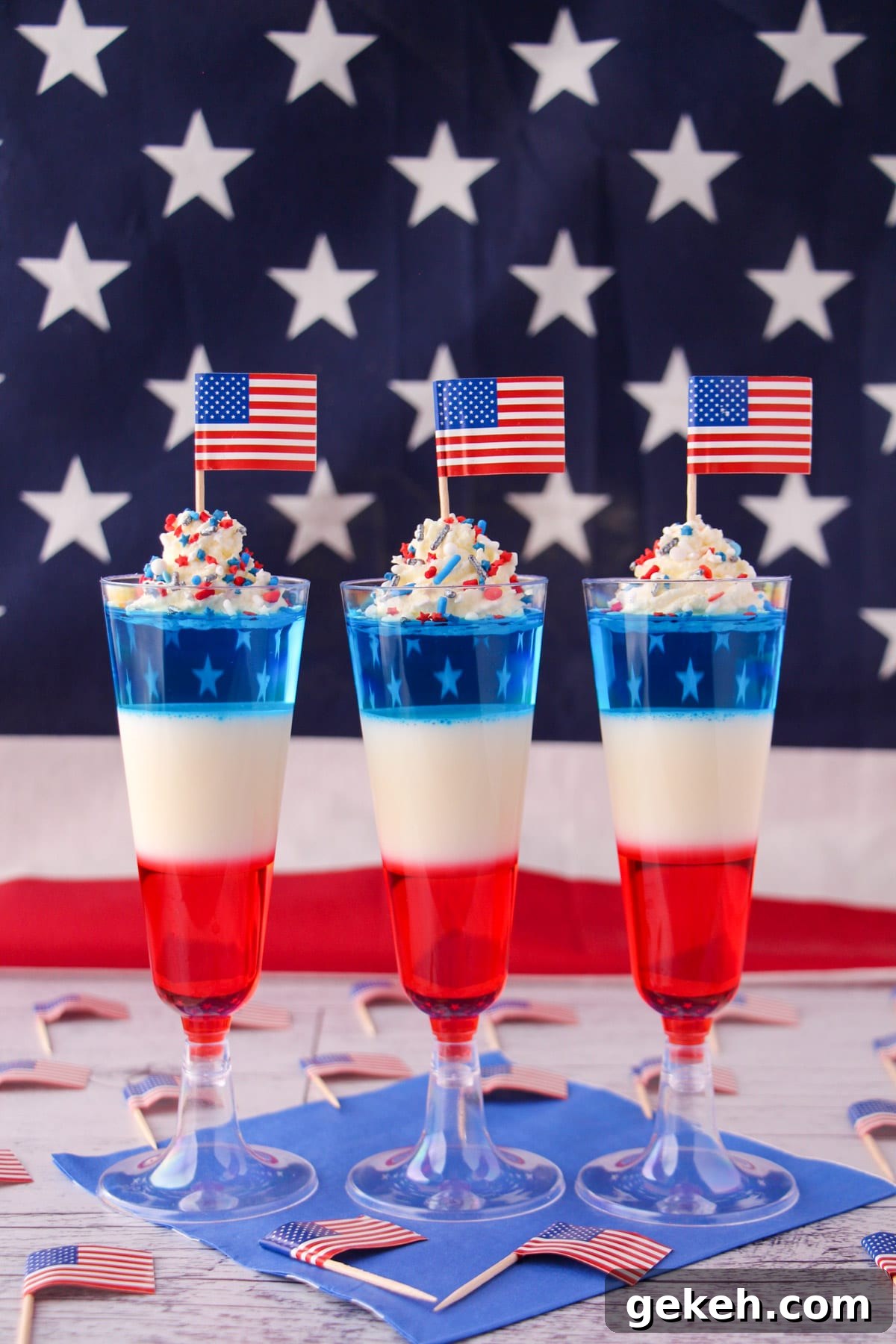 Kid friendly non alcoholic red white and blue jello shots in tall glasses with whipped cream, sprinkles, cocktail stick American flags with American flag in the background.