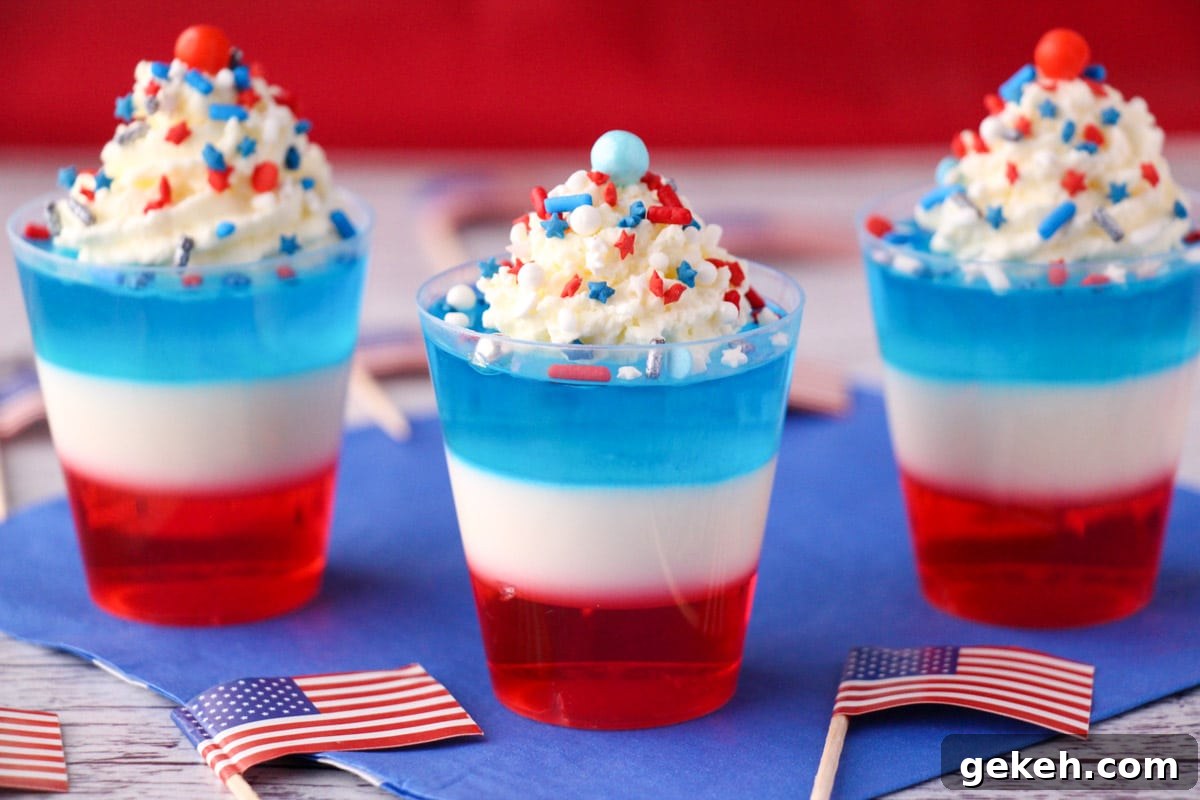 Red white and blue jello shots on blue napkin with whipped cream and sprinkles, surrounded by cocktail stick American flags.