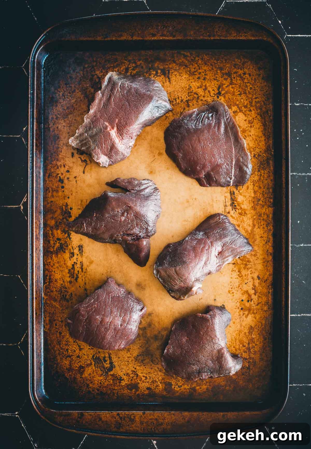 Grilled Elk Steaks: Juicy Elk Steak Recipe and Cooking Tips 3 Six raw elk steaks displayed on a well-used, darkened baking sheet.