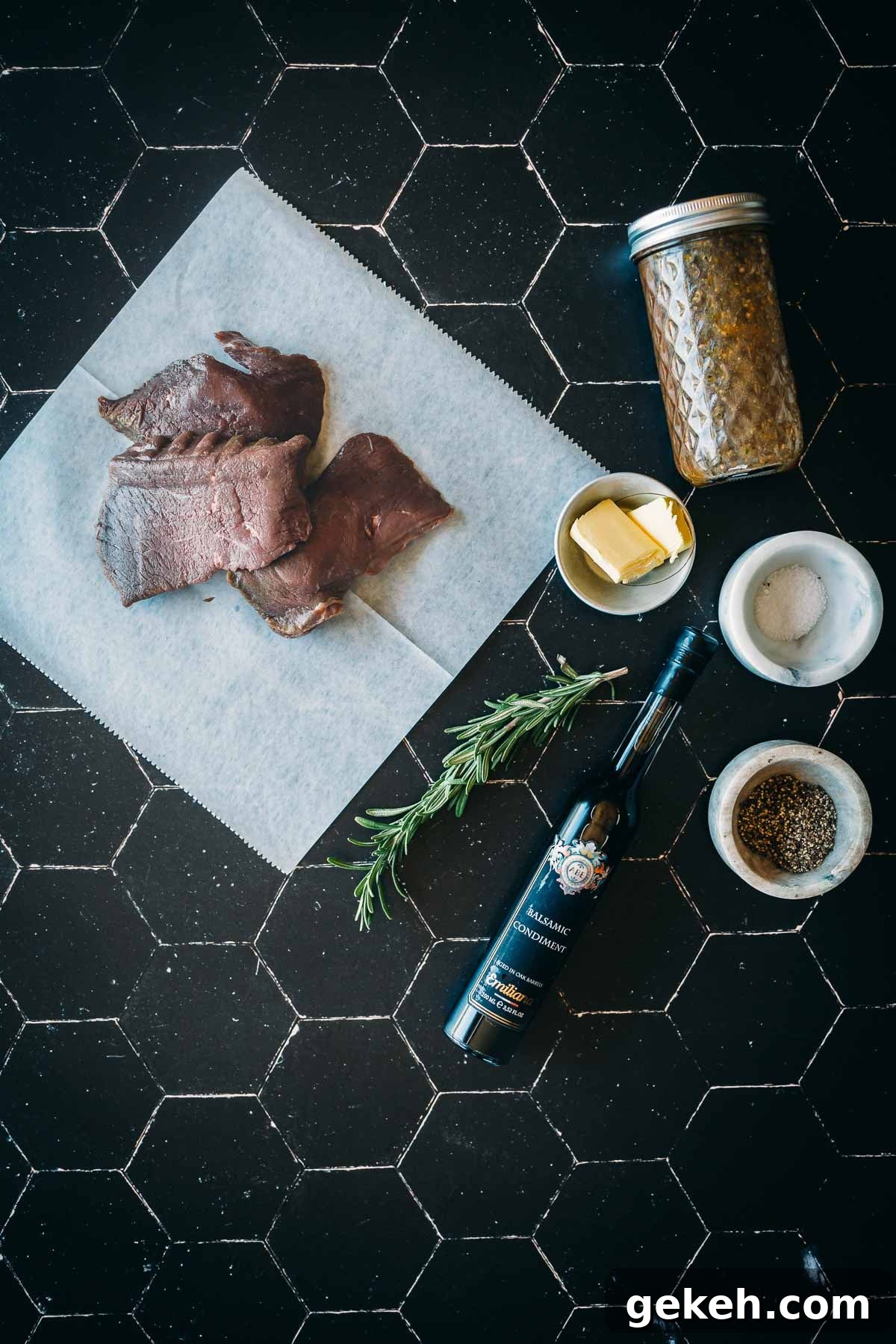 Grilled Elk Steaks: Juicy Elk Steak Recipe and Cooking Tips 4 Raw elk steaks on a parchment paper, jar of seasoning, bowl of butter, rosemary, balsamic vinegar bottle, and bowls of salt and pepper placed on a black tiled surface.