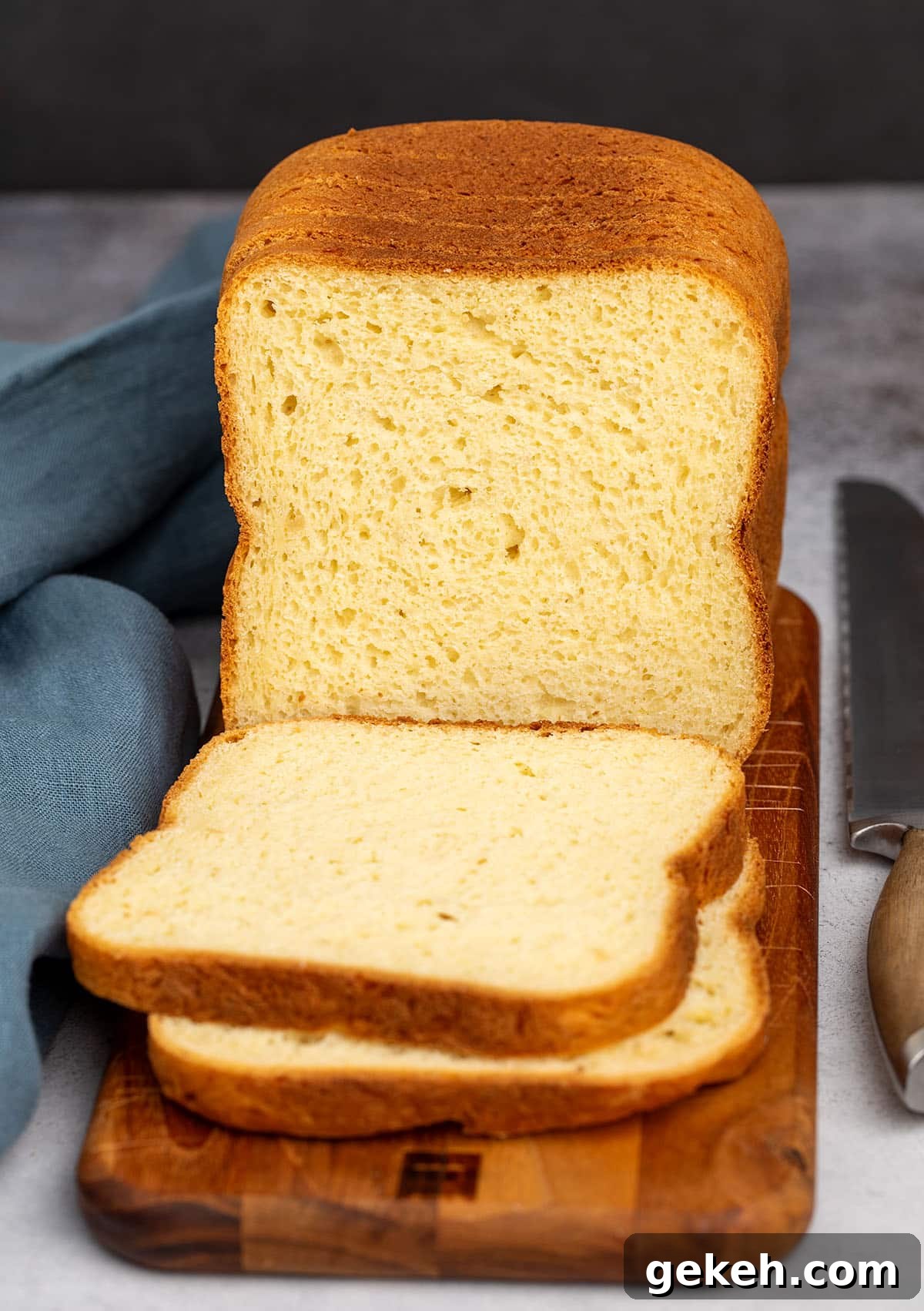 tall partially sliced loaf of bread with a yellow crumb and a brown crust on a narrow dark brown wood cutting board with a blue cloth and metal bread knife with a brown wood handle