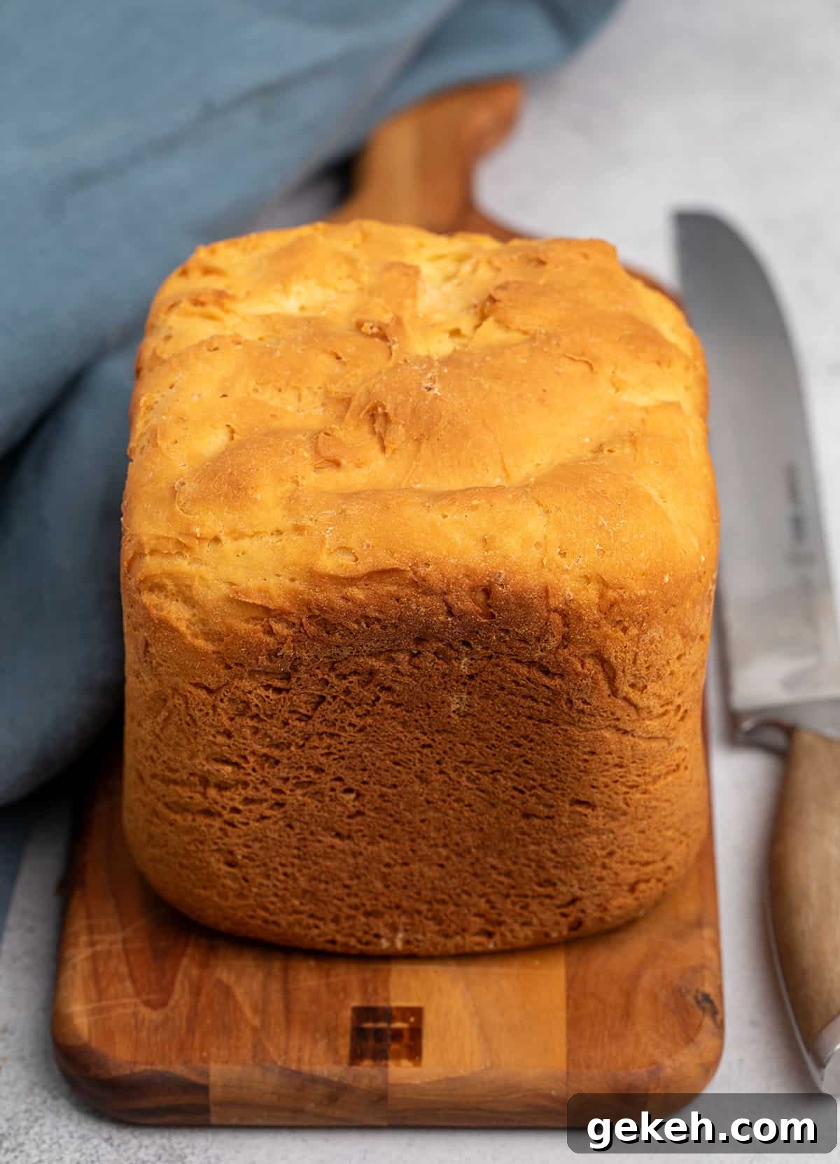 whole brown crusted loaf of bread on the same brown cutting board with blue cloth and bread knife
