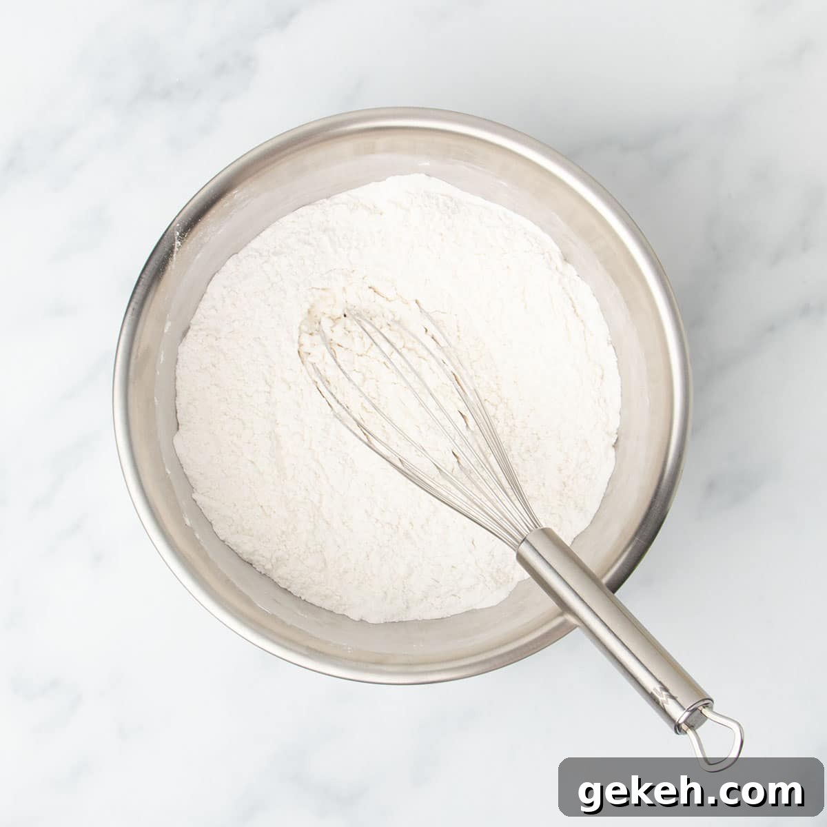 medium round metal mixing bowl with white flour and large metal wire whisk on marble surface