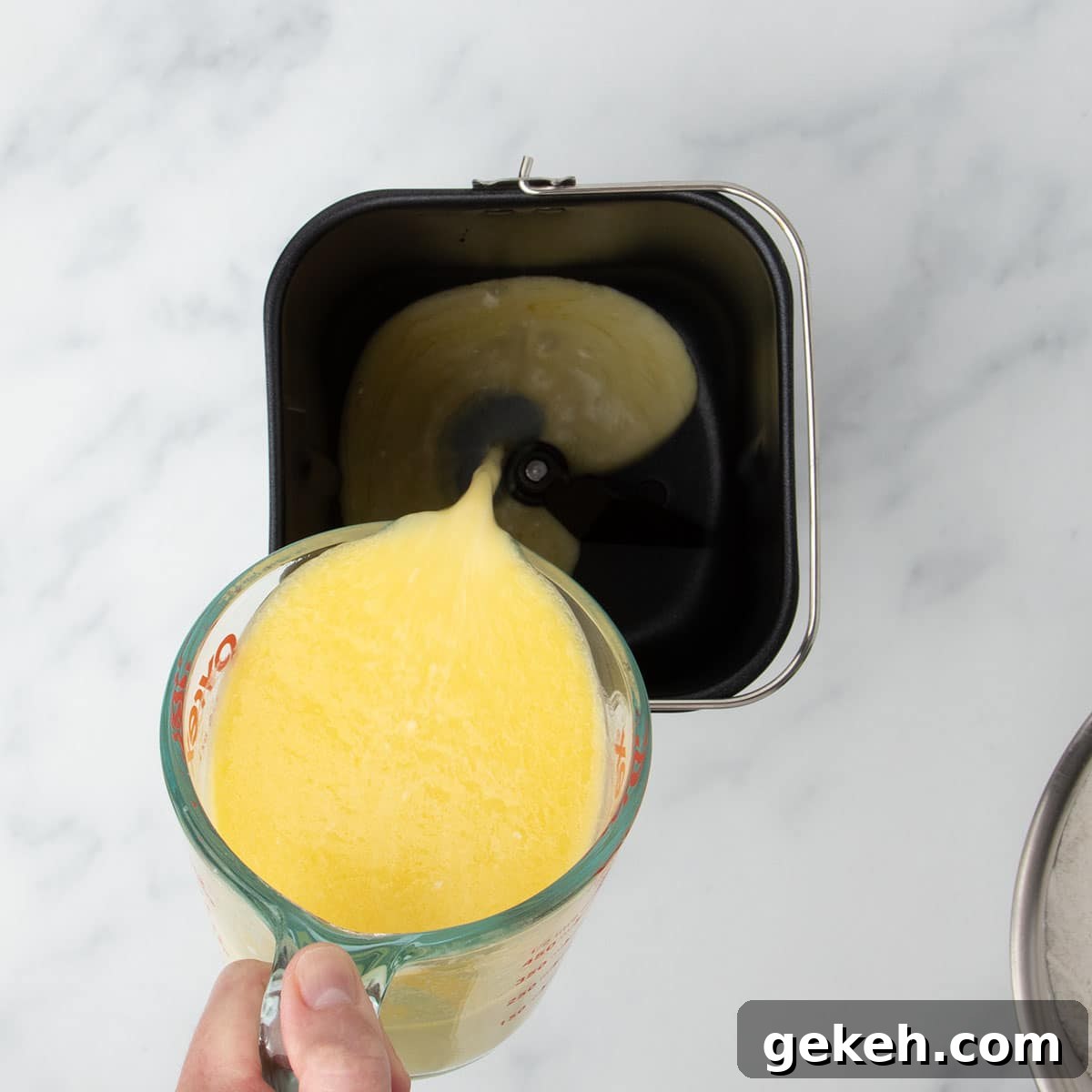 fingers holding 2 cup glass measuring cup pouring yellow mixture into baking pan for bread machine