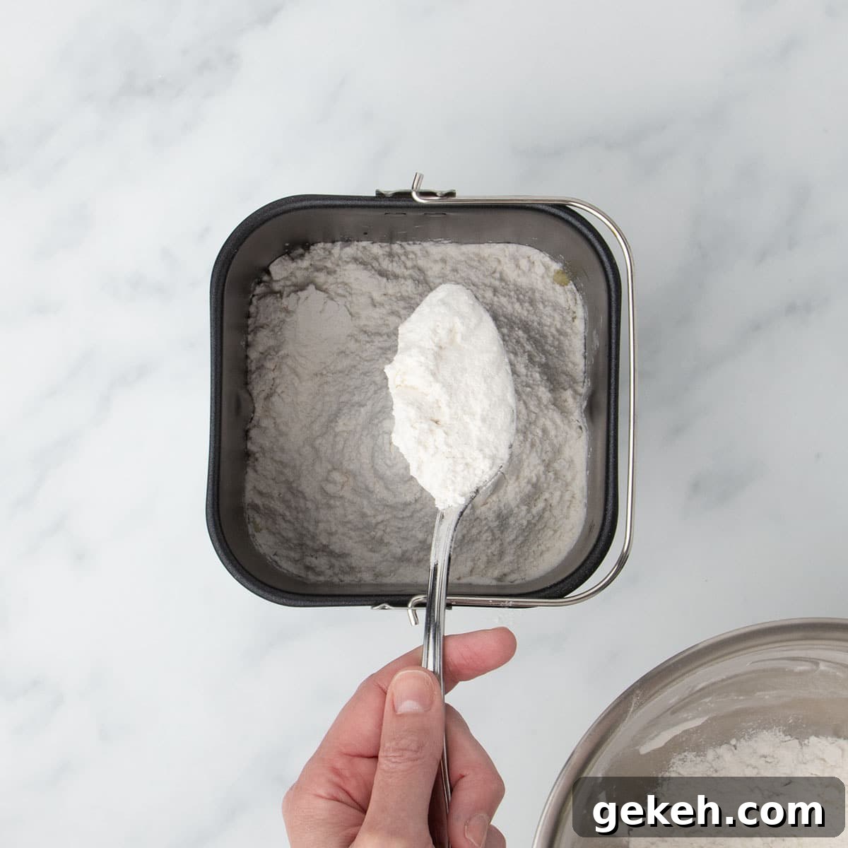 hand holding large metal mixing spoon filled with white flour mixture turning flour over into black bread machine baking pan with more flour