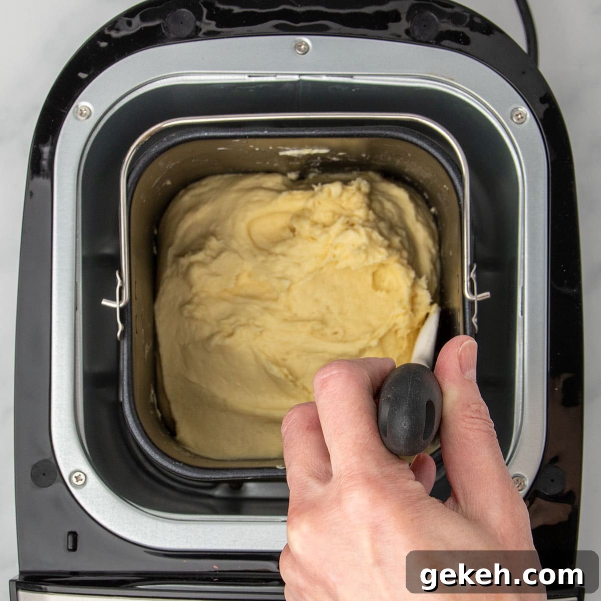 hand holding white silicone spatula with black handle scraping sides of bread machine baking pan with soft raw yellow bread dough